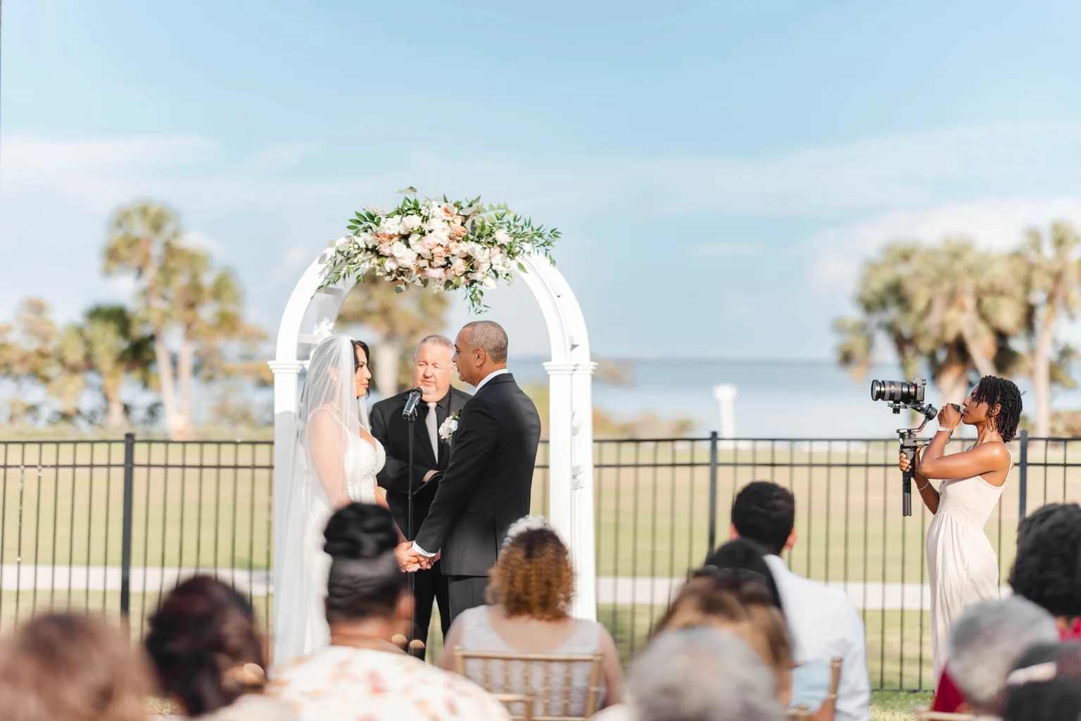 Waterfront wedding ceremony beneath a white floral arch, guests seated in the foreground, with Zxamara Jester filming on a camera gimbal at right.