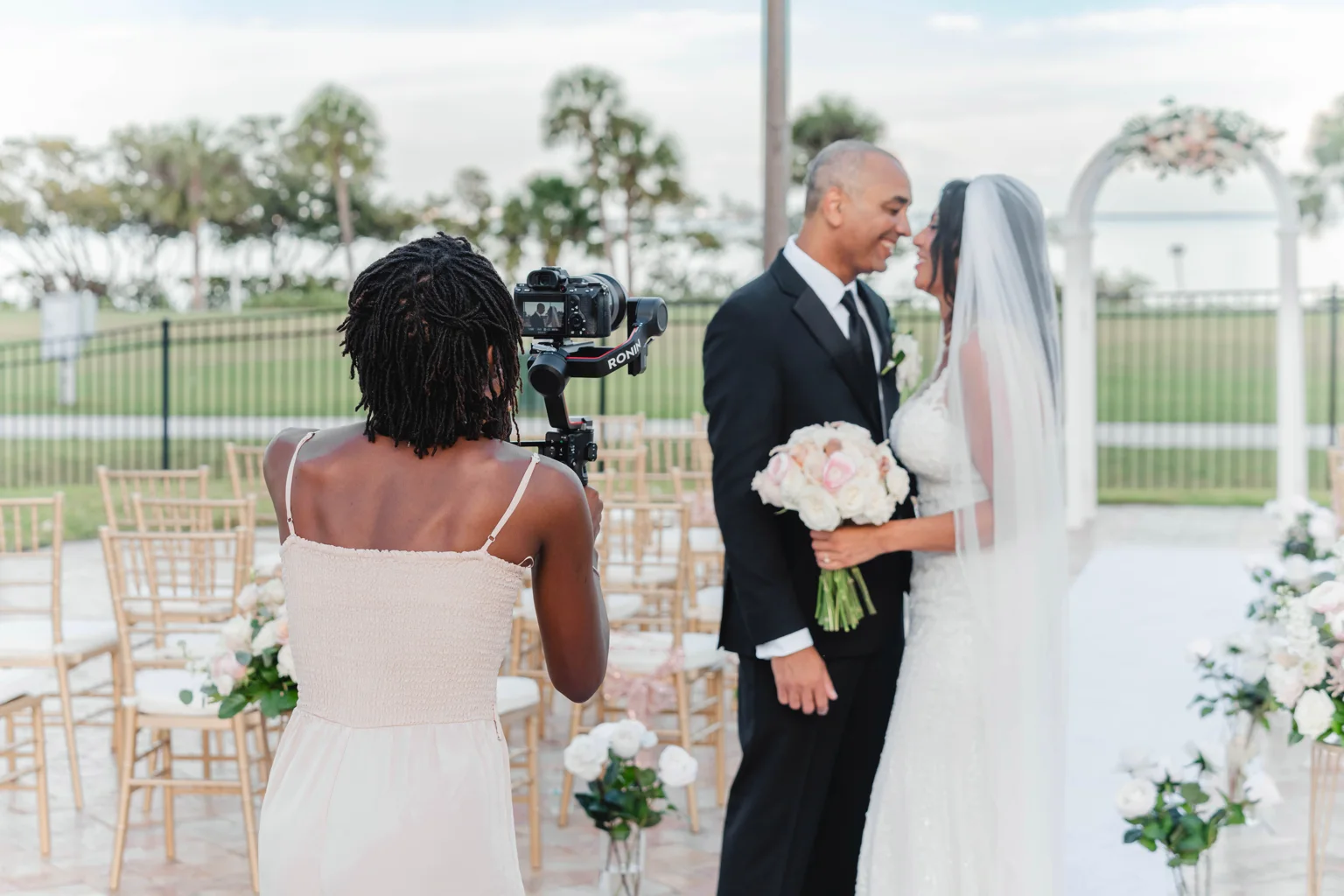 Zxamara Jester filming a smiling bride and groom with a gimbal, gold ceremony chairs and a floral arch behind them.