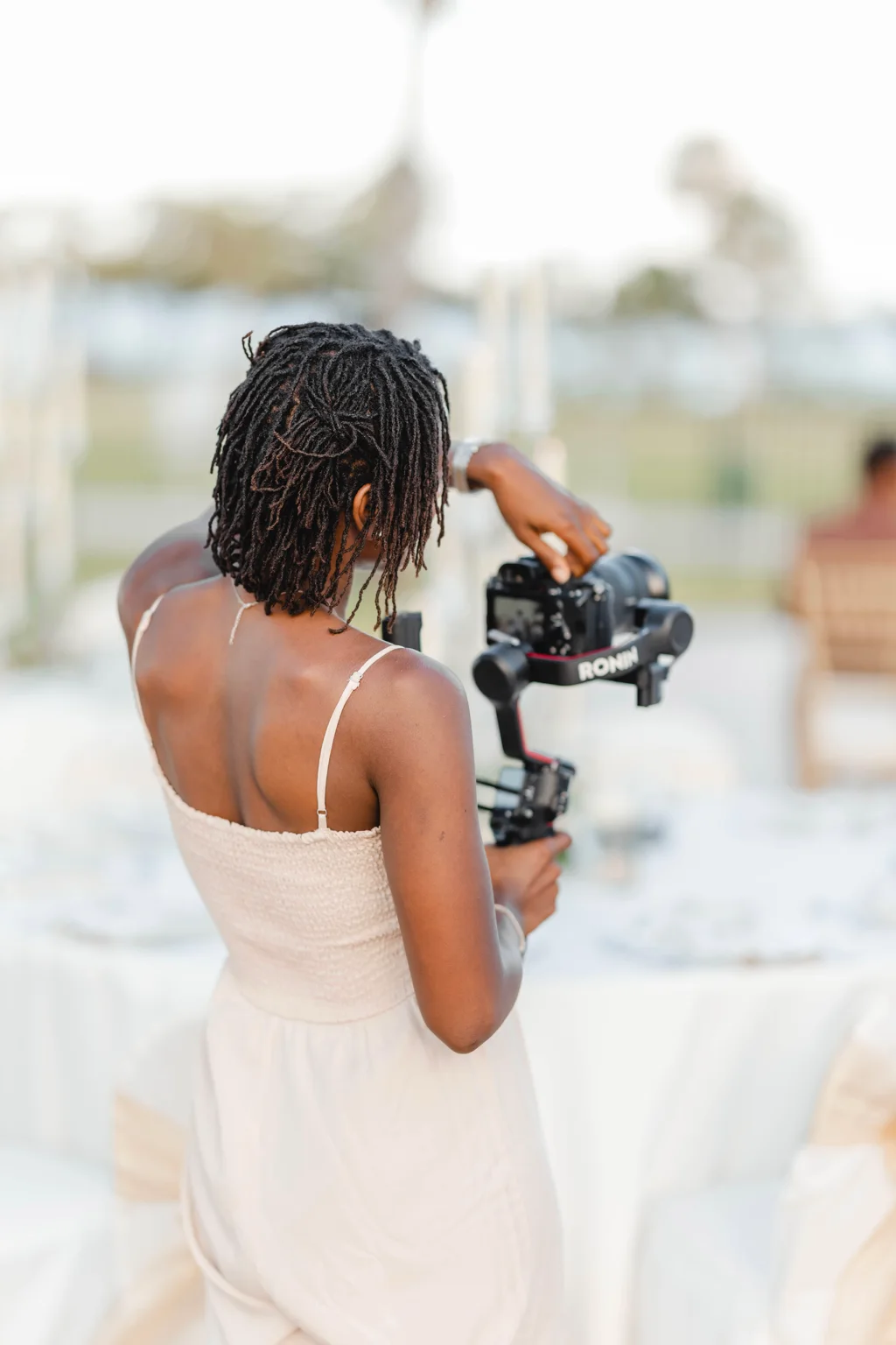 Zxamara Jester seen from behind in a cream outfit, operating a camera on a DJI Ronin gimbal at a wedding reception.
