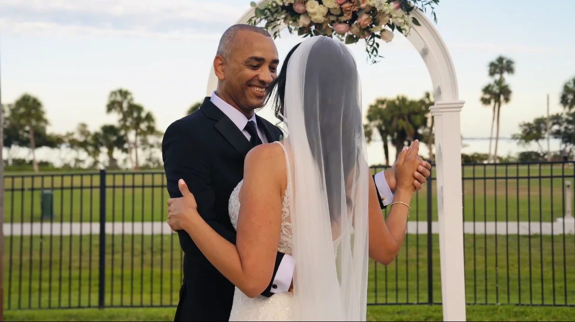 Wedding film still of the groom smiling at the bride beneath a white floral arch, her veil facing the camera.